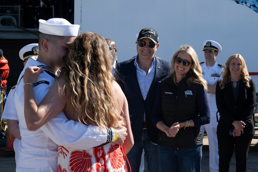 Secretary of War Pete Hegseth and Jennifer Hegseth smile at a sailor who hugs a loved one on a pier.