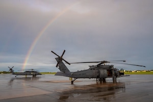 Two helicopters sit on a runway with a rainbow in the sky.
