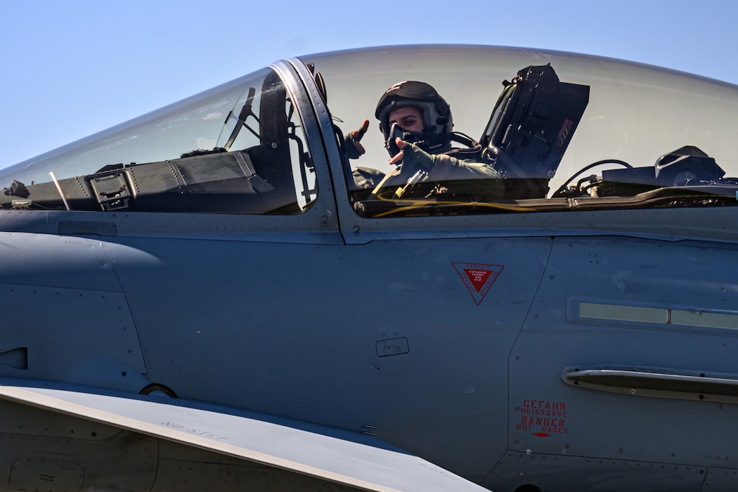 A military fighter pilot points at a camera from inside an aircraft.