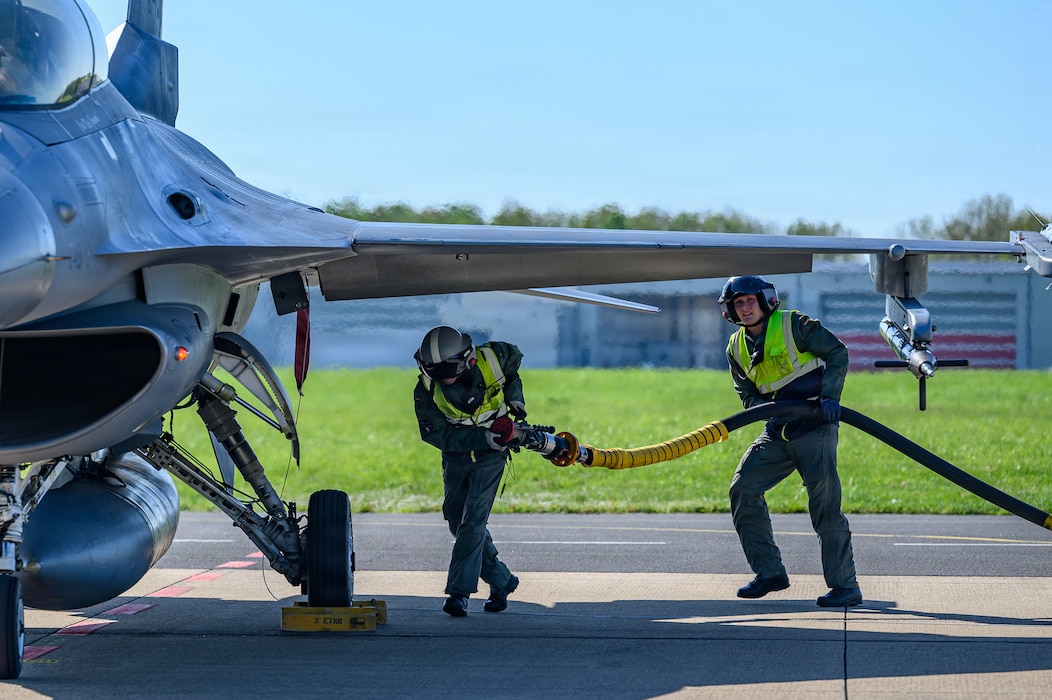 Two people carry a fuel hose under an aircraft.