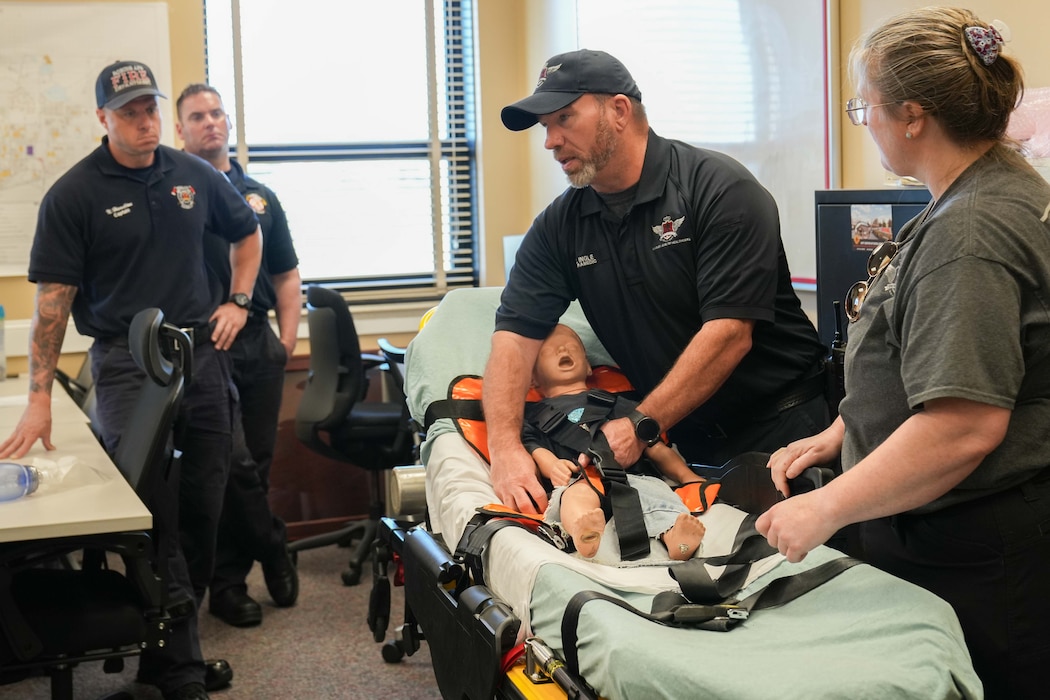 a man straps a child-sized practice dummy into a stretcher three other people watch on