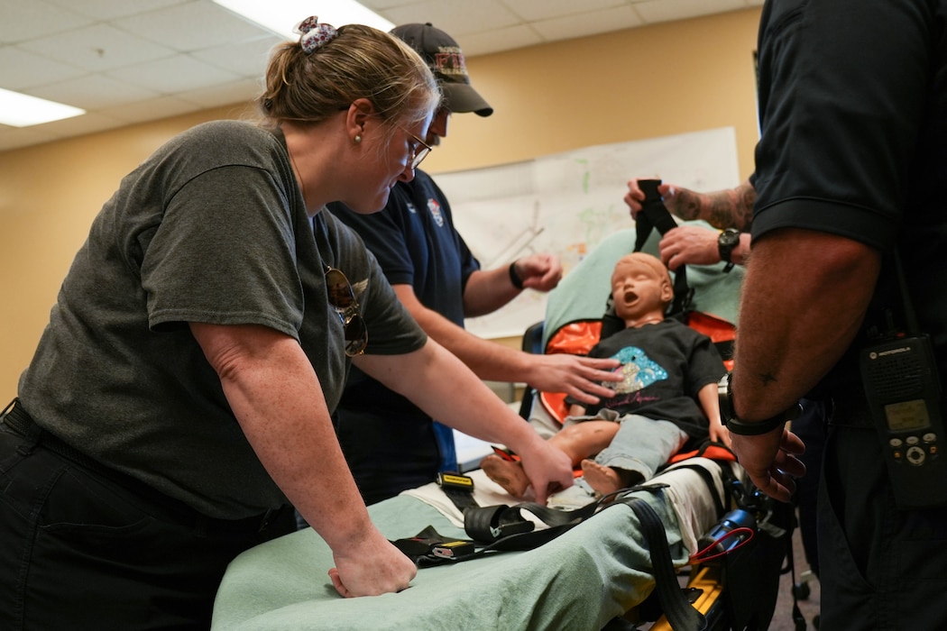 a woman helps to strap a child-sized CPR dummy into a stretcher for safe transport