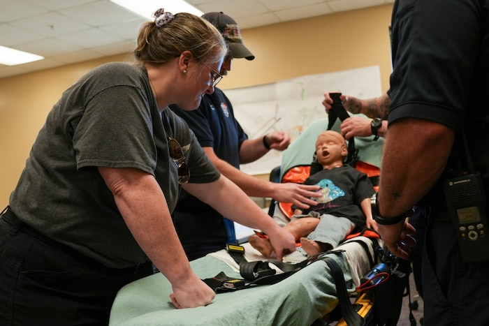 a woman helps to strap a child-sized CPR dummy into a stretcher for safe transport