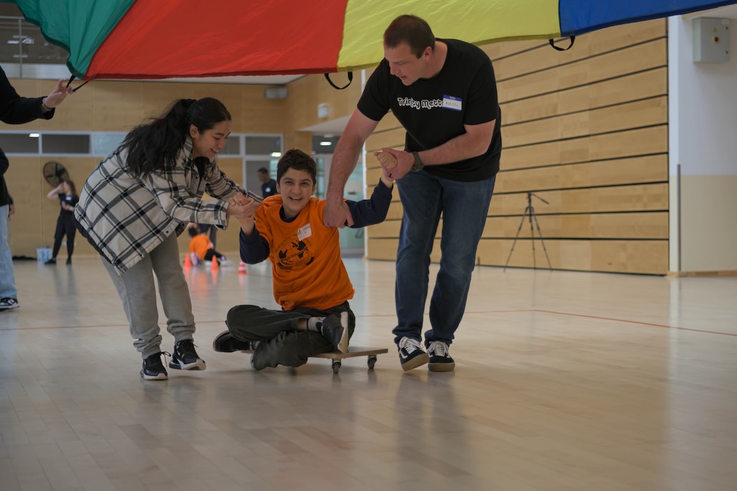 Two people pull a child on a scooter through a gym.