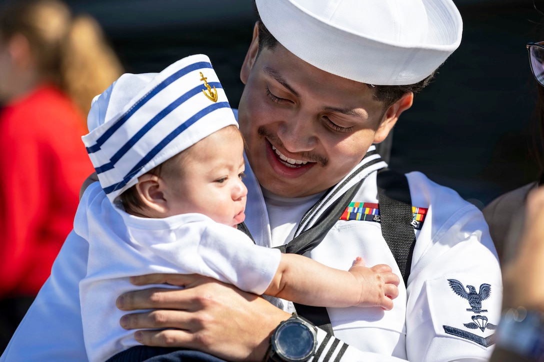 A close-up of a sailor in uniform holding a newborn wearing a sailor's hat.