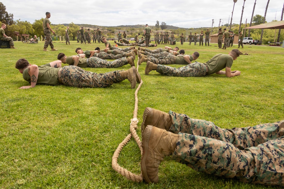 Marines lie on a field on opposite sides of a rope as fellow Marines train in the background.