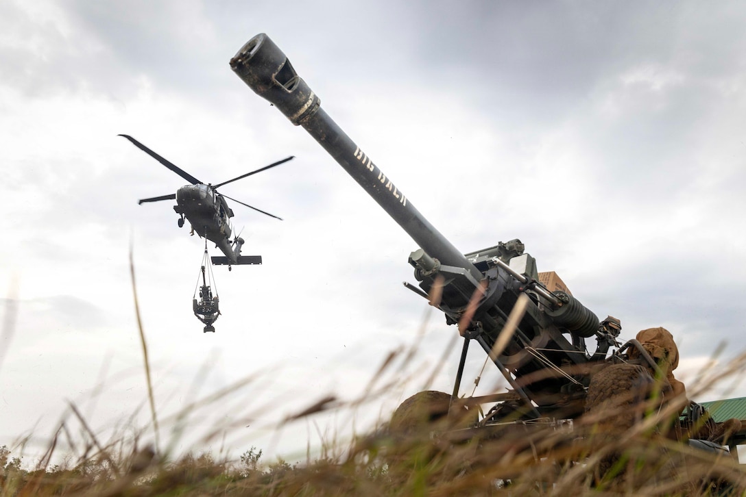 An airborne helicopter sling-loads a weapon in a gray sky as another weapon sits in the grass in the foreground.