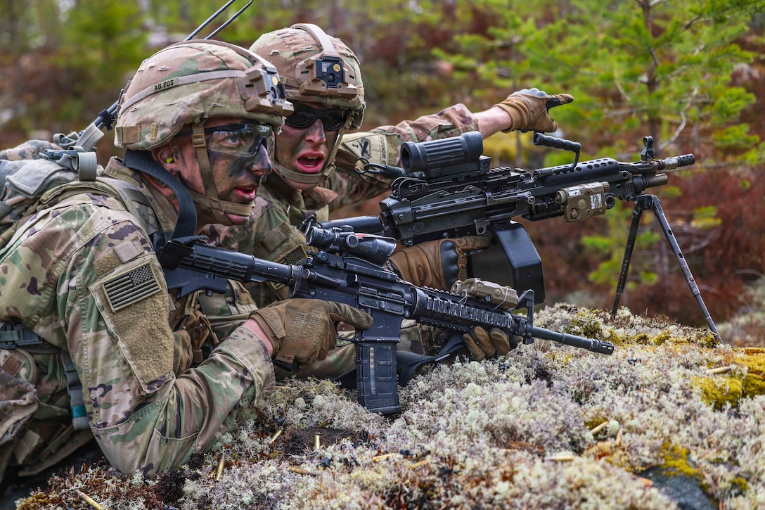 Two soldiers hold machine guns while lying on their stomachs on a hill. One soldier points with his left arm while they look toward the right.
