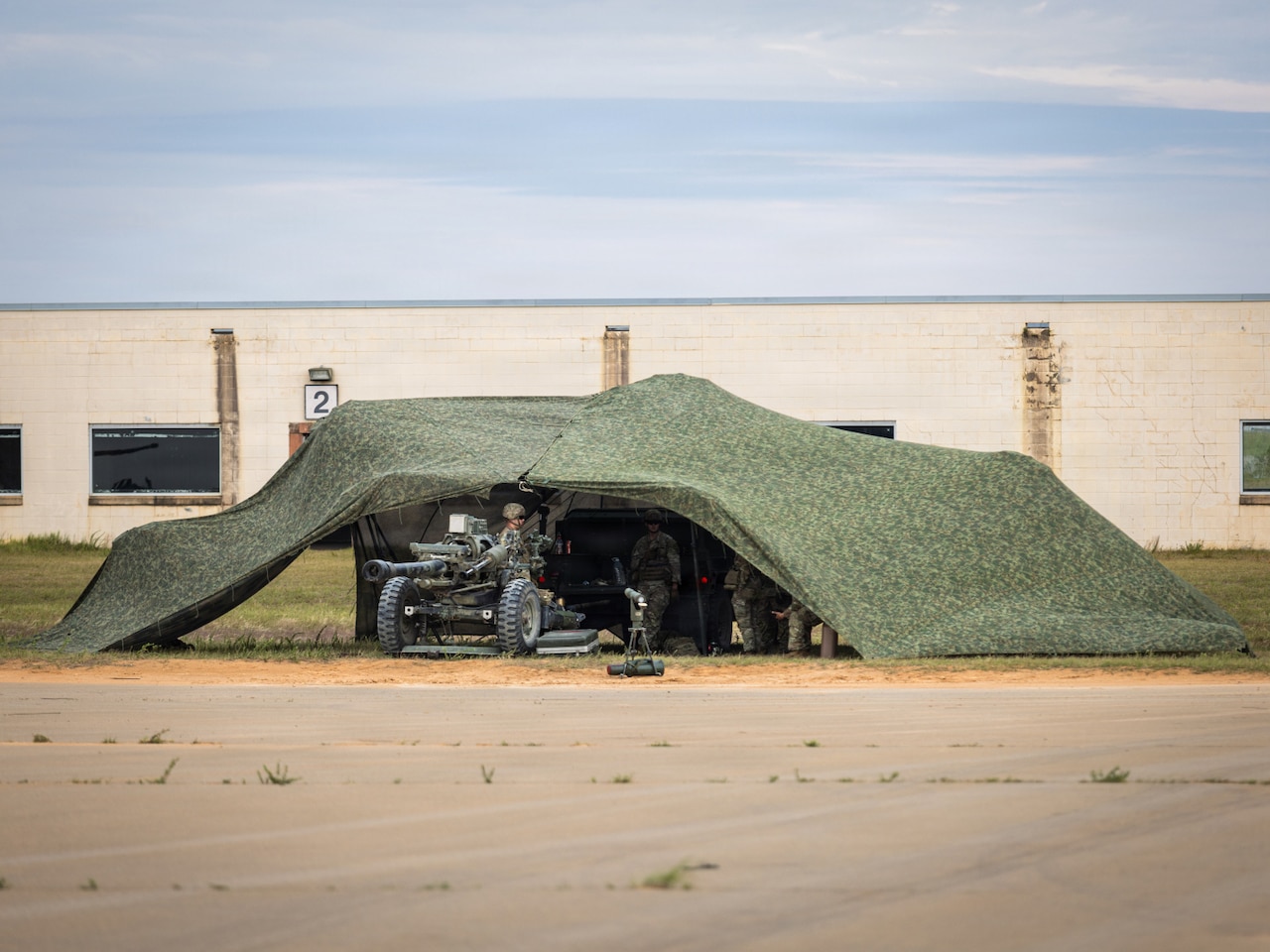 A group of people wearing camouflage military uniforms stand under a camouflage military tent. One of the people stands near a military tank just outside of the tent.