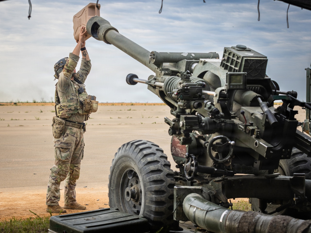 A soldier wearing a military camouflage uniform holds a container and pours liquid into the barrel of the gun of a military tank.