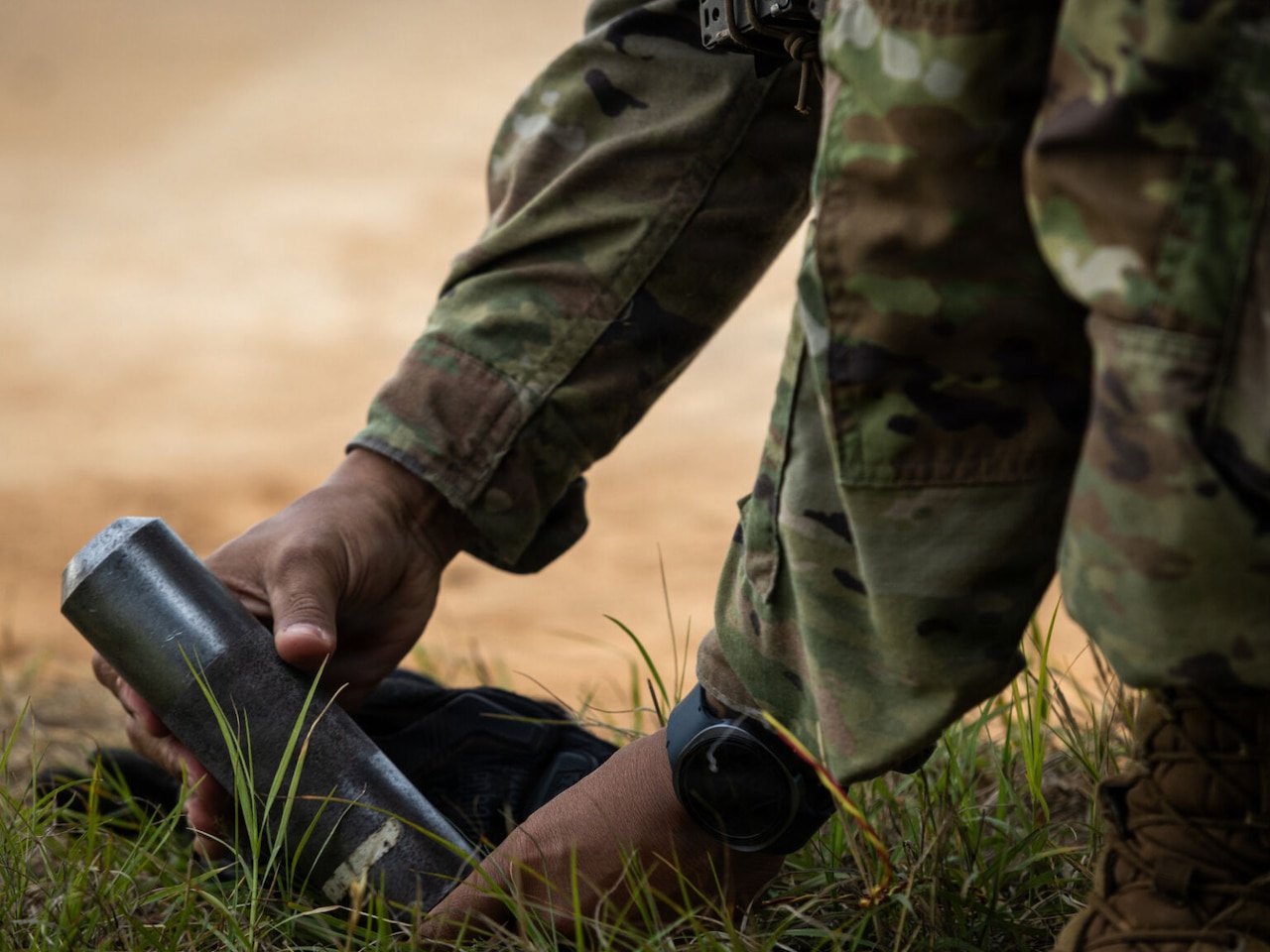 A person wearing a camouflage military uniform picks up an artillery round from the grassy ground.