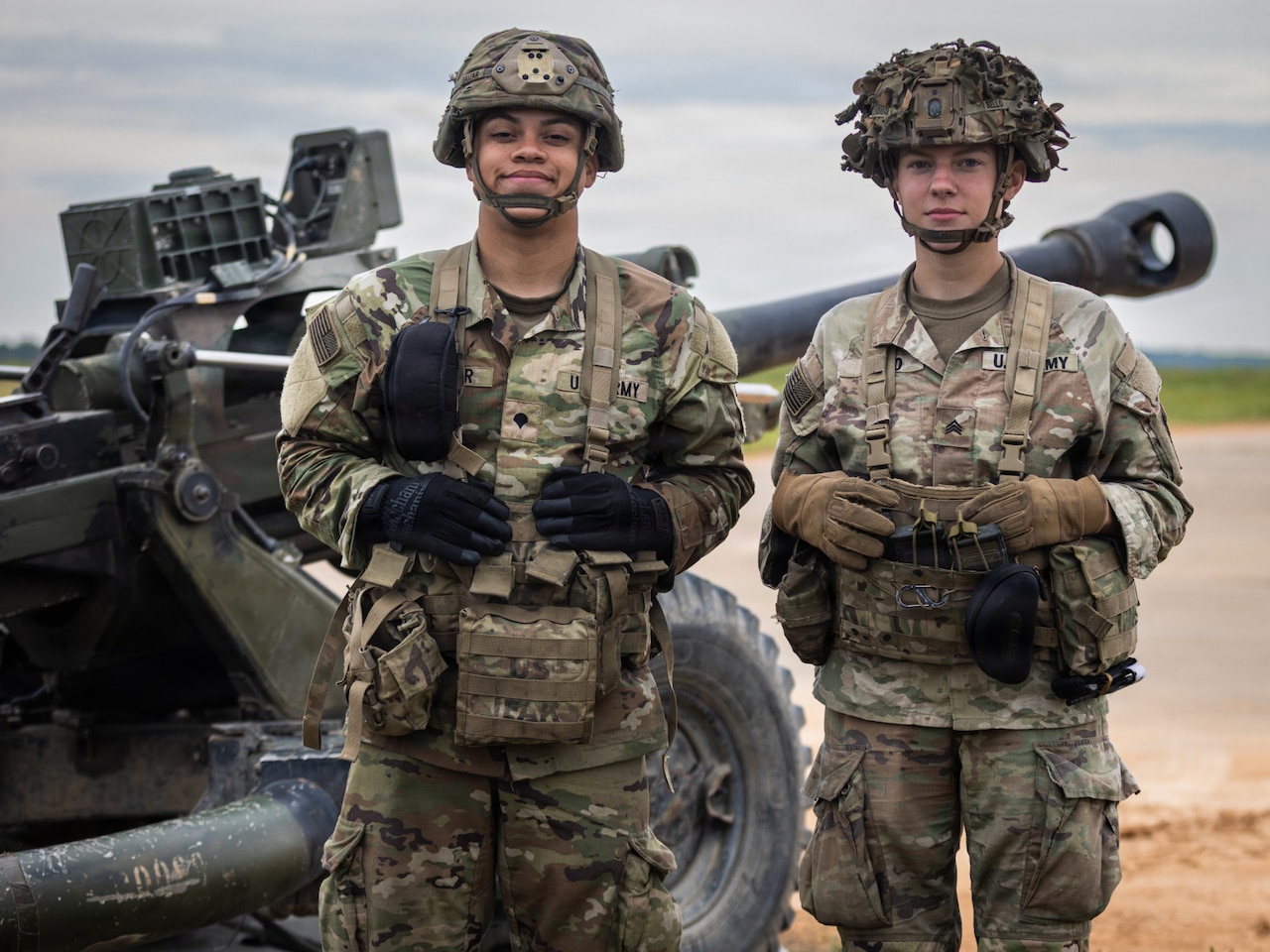 A man and a woman wearing camouflage military uniforms stand and smile as they pose for a photo. A large military tank is behind them.