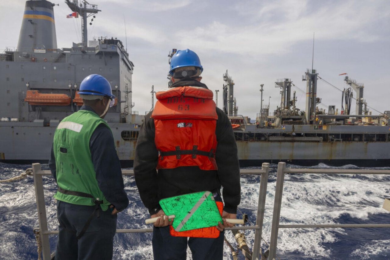 Two people in military uniforms, hard hats and reflective vests stand at a ship's rail, looking across the water at another large vessel.