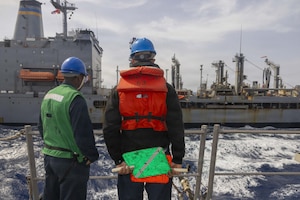 Two people in military uniforms, hard hats and reflective vests stand at a ship's rail, looking across the water at another large vessel.
