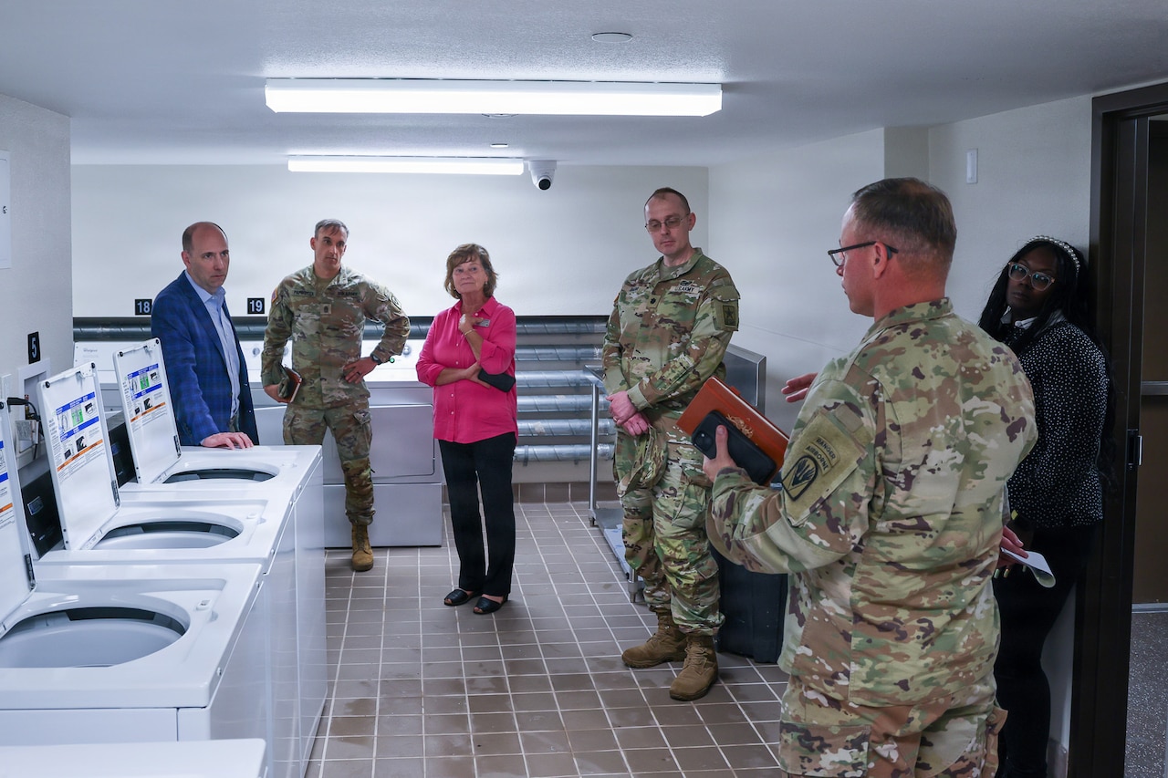 Six people, some in camouflage military uniforms and others in business attire, stand in a laundry room listening to a man speak.