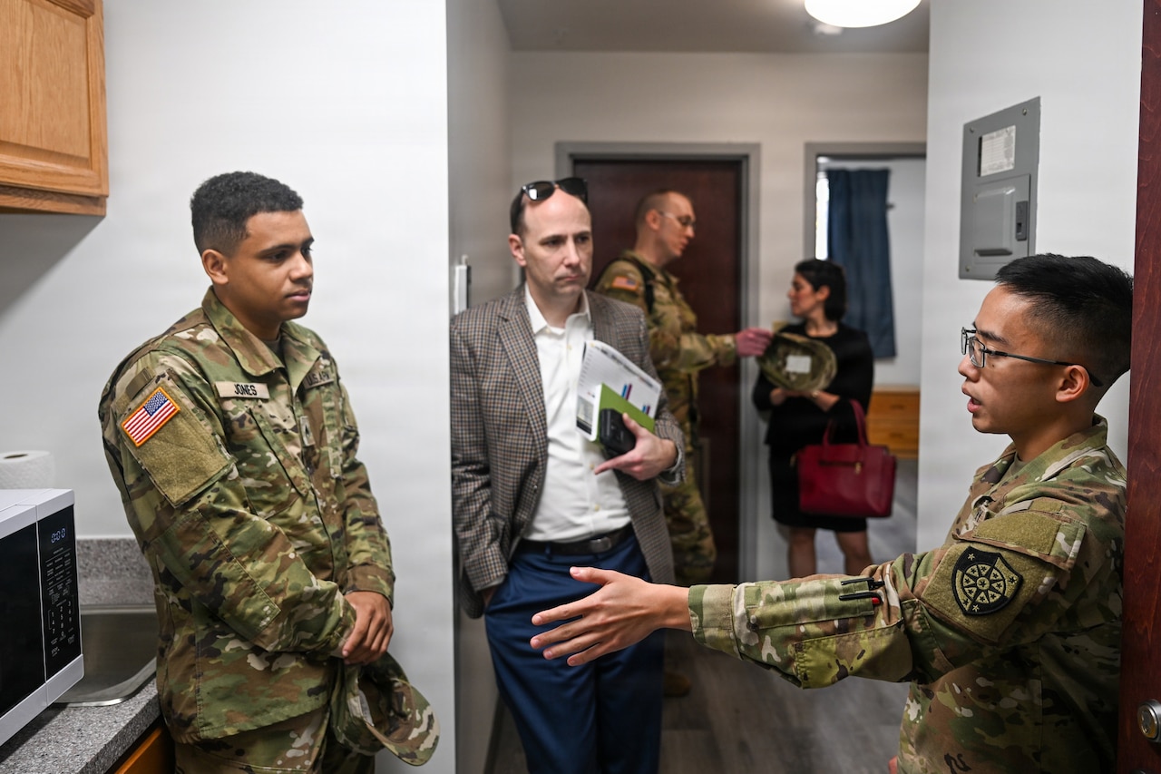 A man wearing business attire listens while two people in camouflage military uniforms speak inside a small kitchen.  In the background, a man and a woman have a conversation.