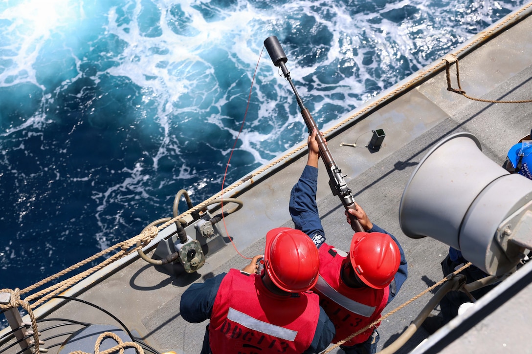 Two sailors in red hard hats and red vests stand on a ship’s deck. One holds a tool with a red string hanging out of it over the edge of the ship. Turquoise water is in the background.