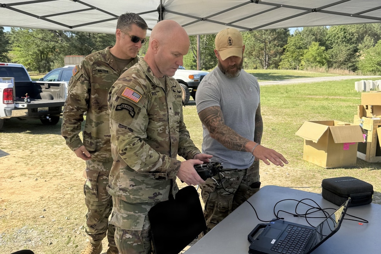 A man wearing casual attire instructs a man in a camouflage military uniform on how to use a drone remote control as another man wearing a camouflage military uniform observes. They are standing outside under a pop-up tent in a field, looking at a computer sitting on a table.