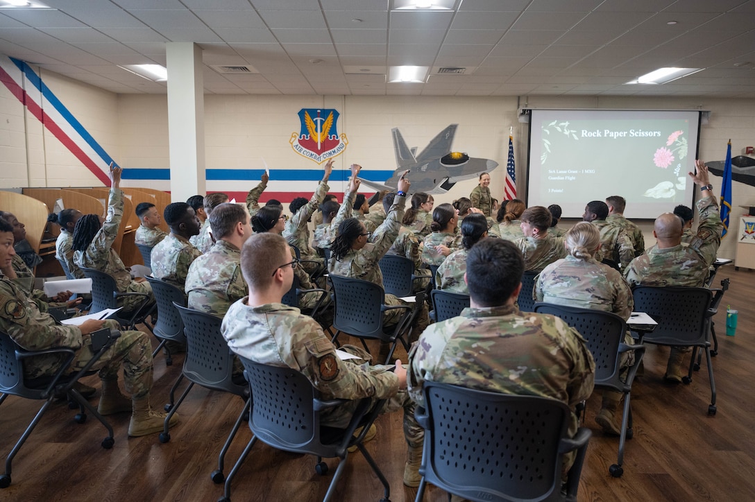 A group of U.S. Airmen raise their hands amongst a classroom of peers all sitting in chairs.