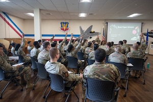 A group of U.S. Airmen raise their hands amongst a classroom of peers all sitting in chairs.