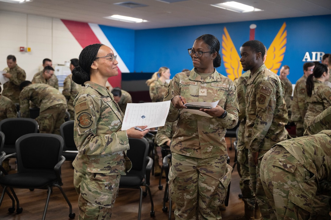Two female U.S. Airmen smile while talking amongst a classroom of peers.