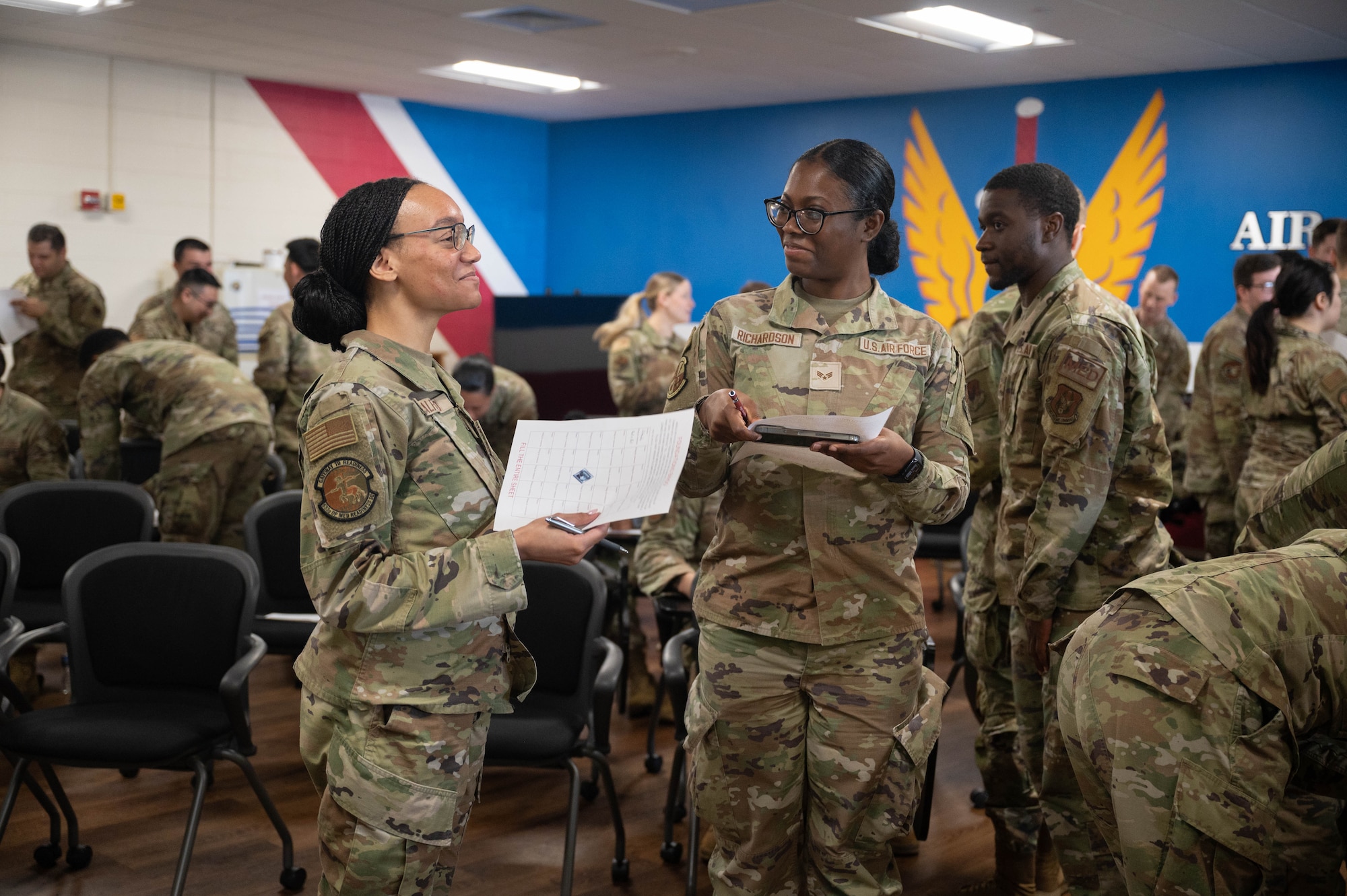 Two female U.S. Airmen smile while talking amongst a classroom of peers.