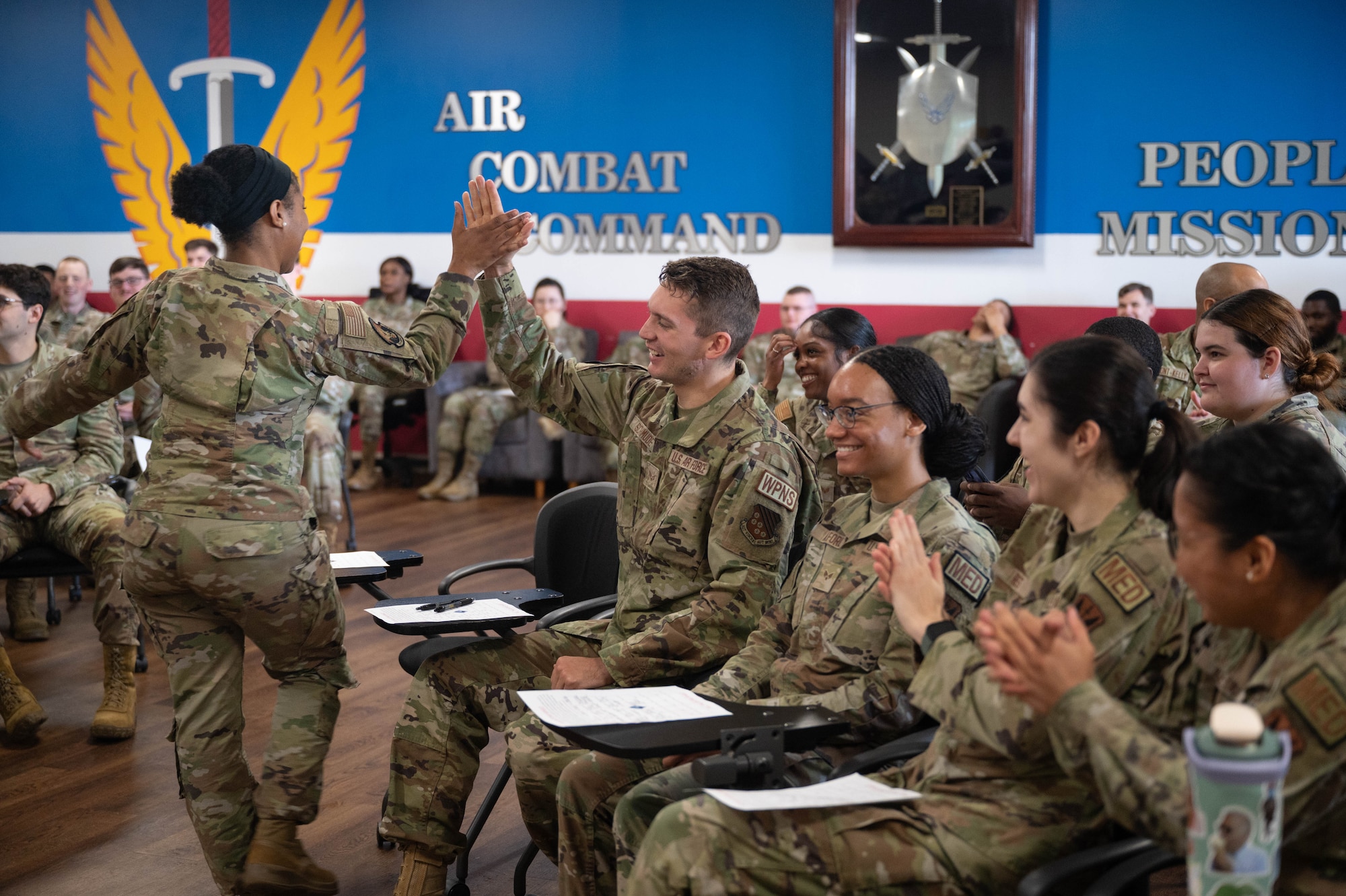 Two U.S. Airmen high-five each other among a classroom of peers sitting at chair desks.