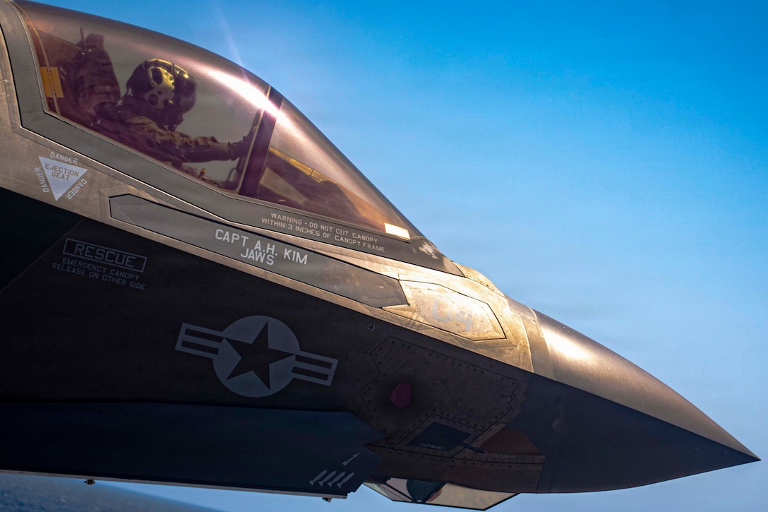 Sunshine reflects off the top of a fighter jet with a star outlined in white paint on its side and a pilot inside. A blue sky and a blue sea are in the background.