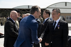 A person in formal civilian attire shakes hands with a person wearing a suit, with a medal attached to a purple ribbon on the blazer, during daytime outdoors; other people are standing in the background.