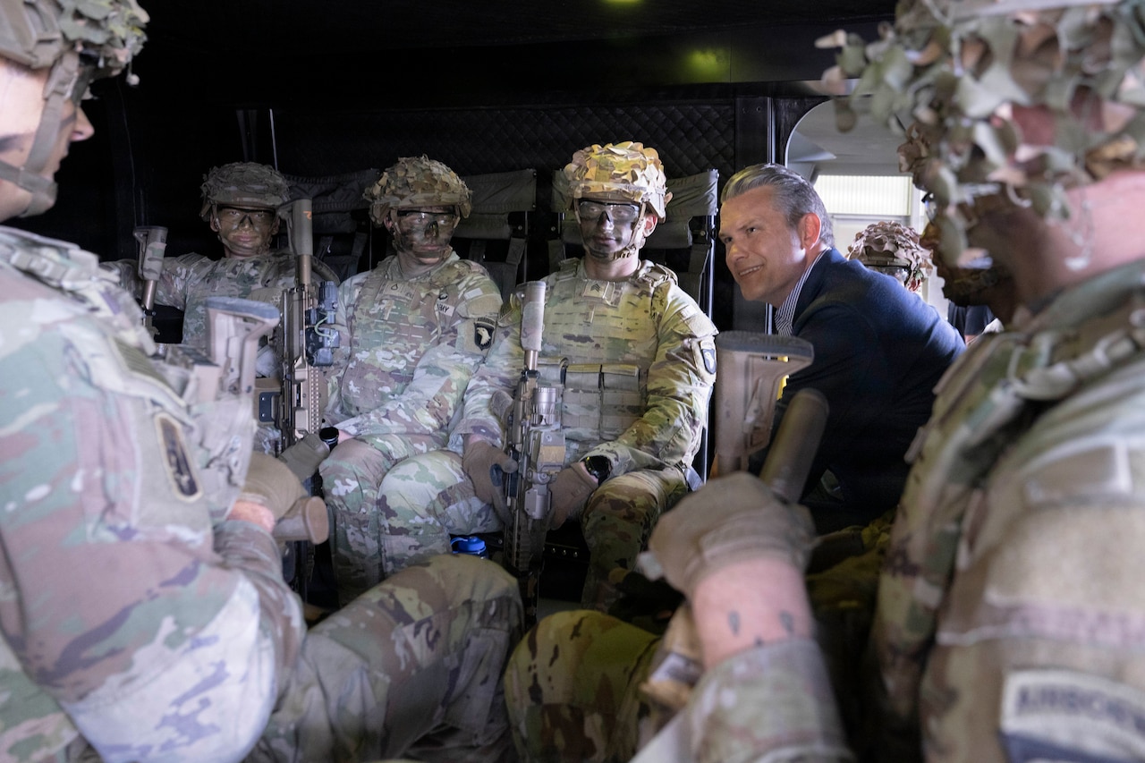 A person in formal civilian attire sits with soldiers in camouflage tactical gear inside a small enclosure.