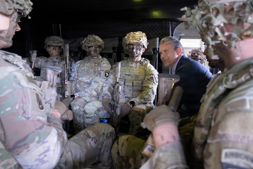 A person in formal civilian attire sits with soldiers in camouflage tactical gear inside a small enclosure.