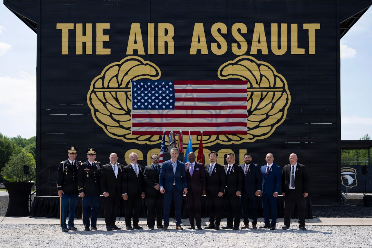 Seven people in business attire and two people in officer uniform pose for a photo with Secretary of War Pete Hegseth, with an American flag on a wooden wall behind them outside, painted wings behind the flag and the words The Air Assault above the flag in yellow.