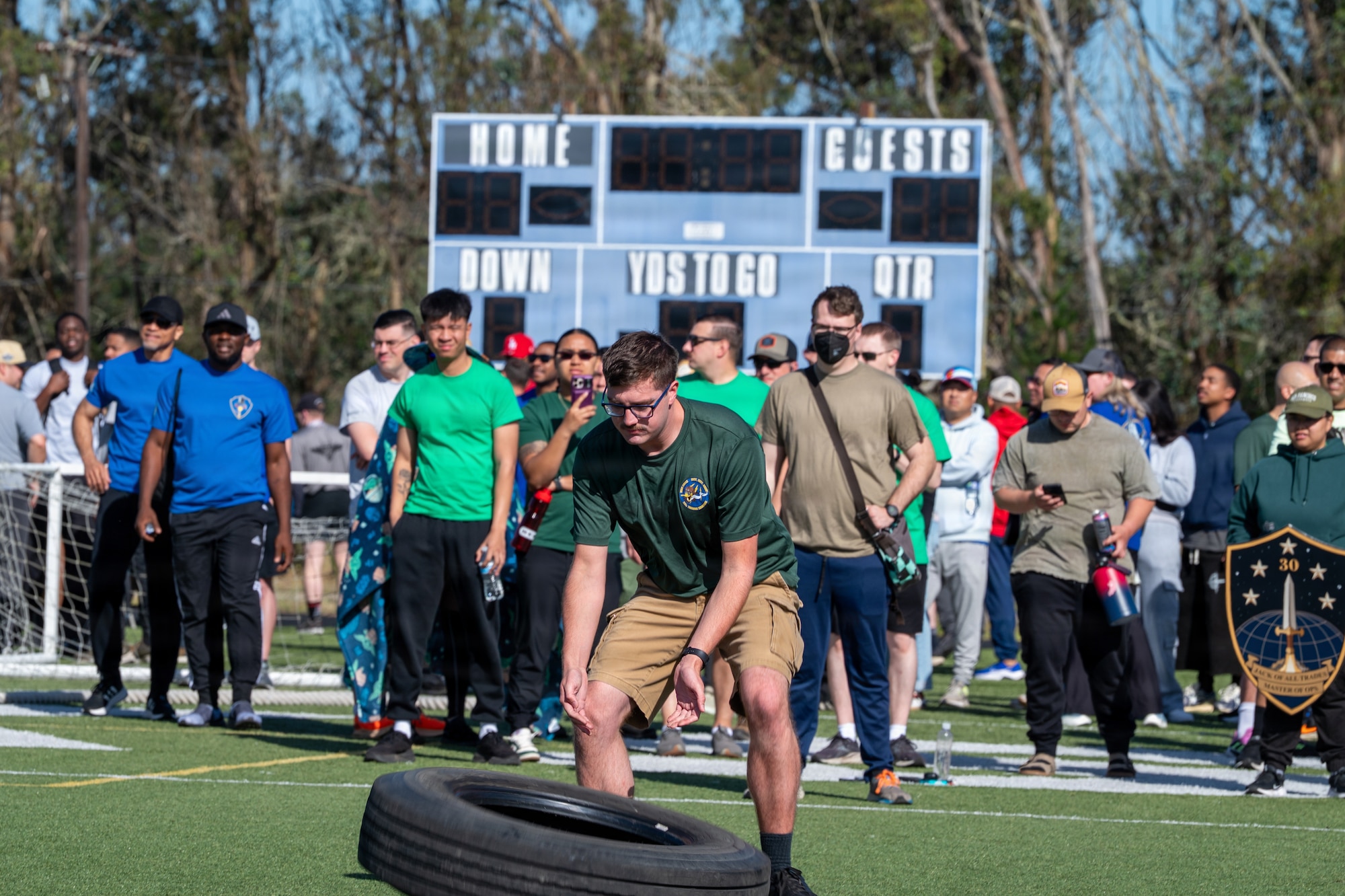 A person moves a tire outside on a turf field as a crowd of people watches.
