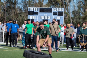 A person moves a tire outside on a turf field as a crowd of people watches.
