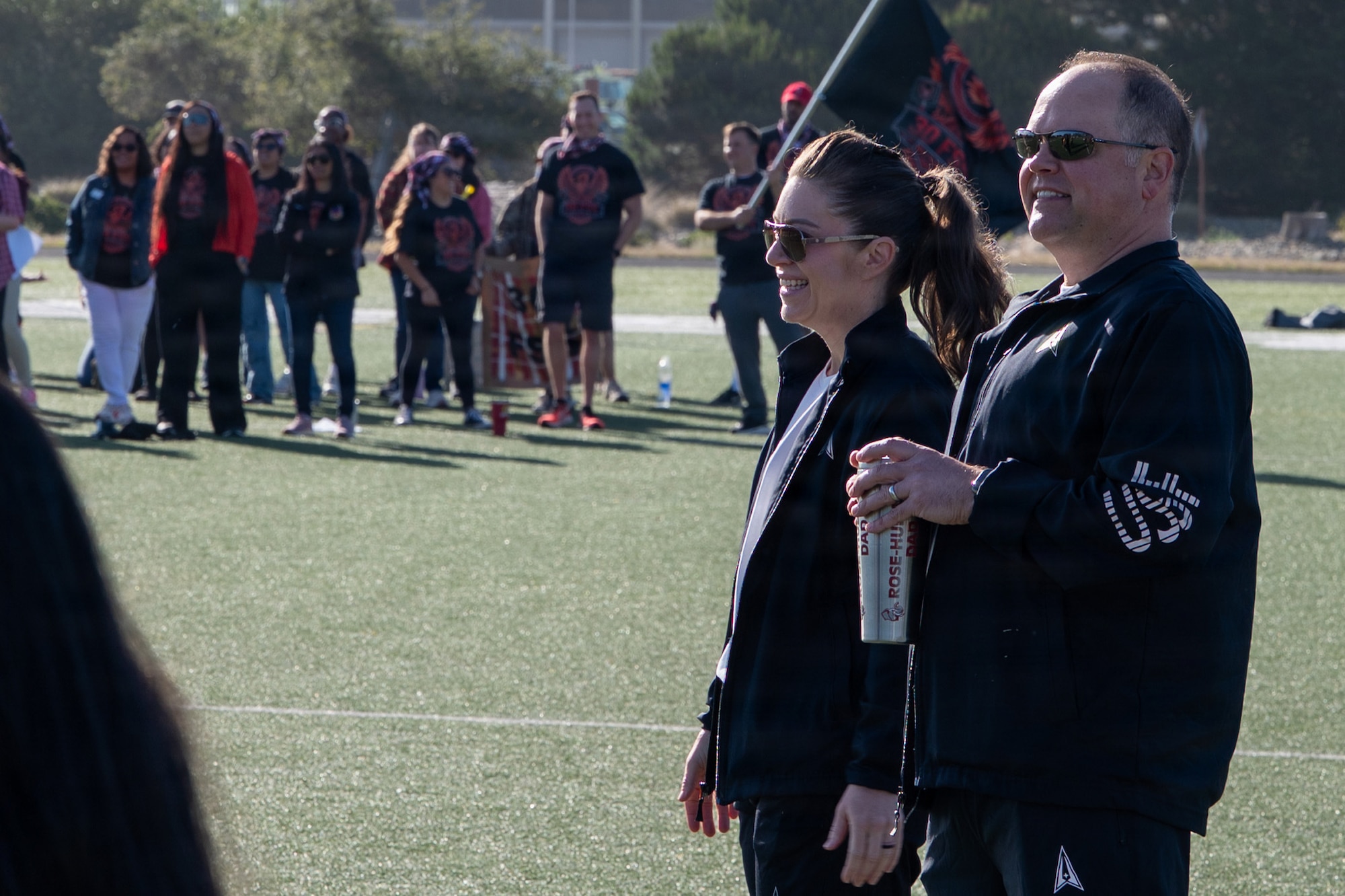 Two people stand next to each other and smile as they look out outside on a turf field.