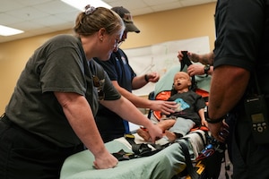 a woman helps to strap a child-sized CPR dummy into a stretcher for safe transport