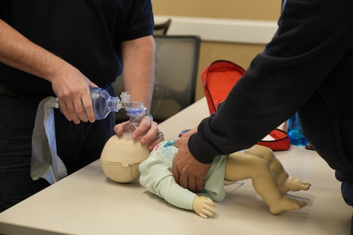 two people perform CPR and bagged rescue breaths on an infant-sized CPR dummy