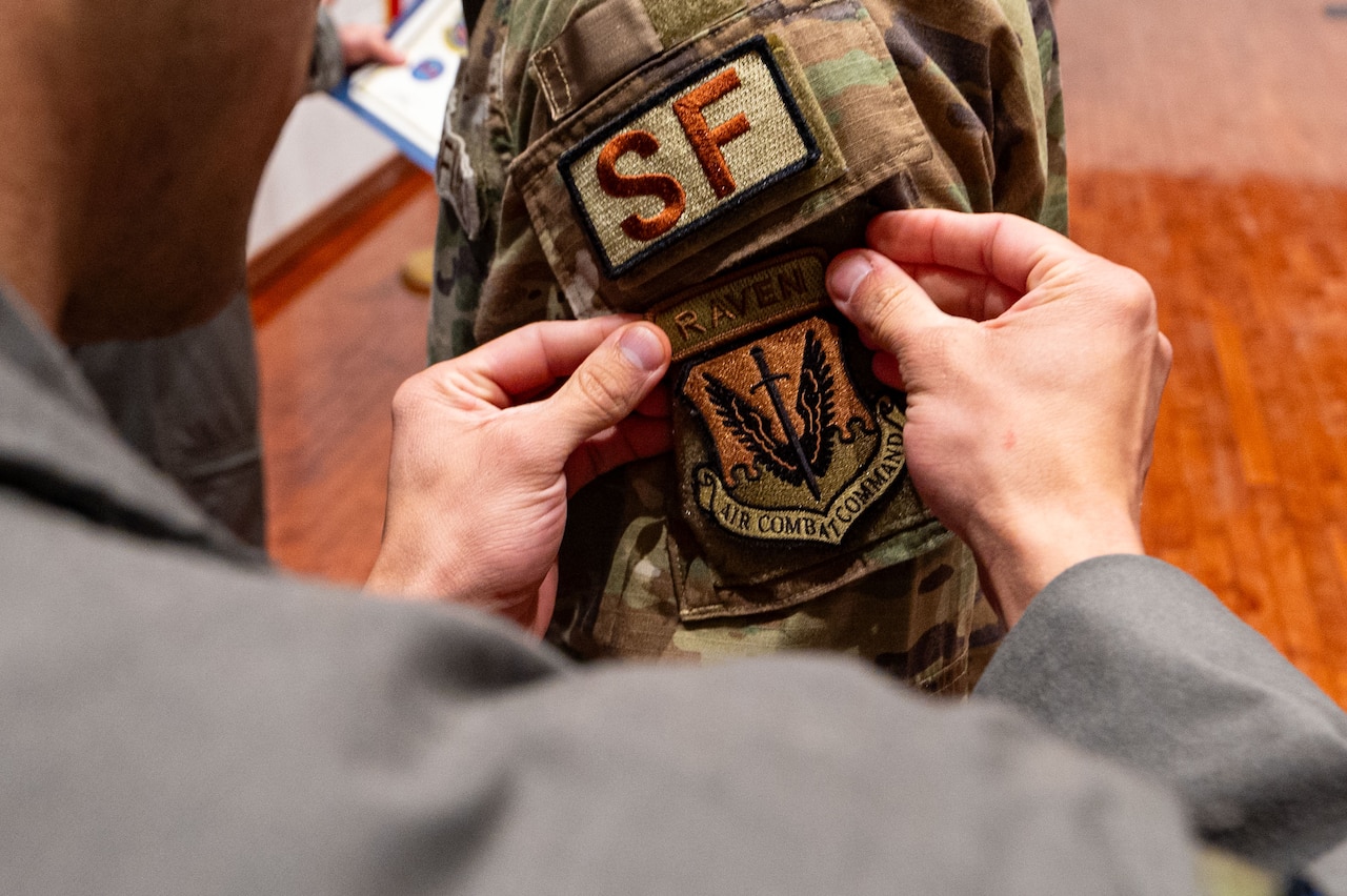 A close-up of a person attaching a patch to the sleeve of another person's camouflage military uniform. The patch reads, "Raven, Air Combat Command."