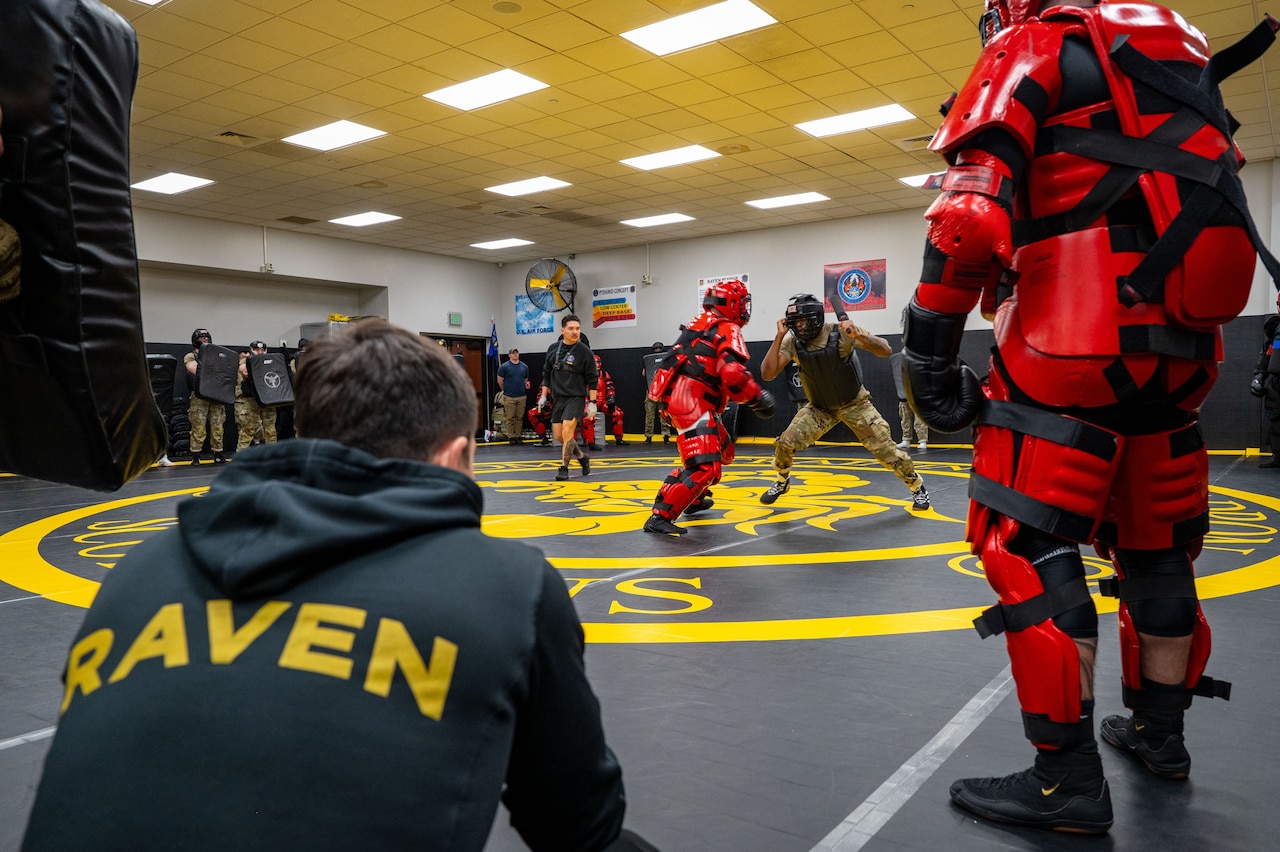 Two people in protective gear practice self-defense techniques on a wrestling mat while dozens of other people in similar, casual and military attire watch from around the room. There is a man crouched in the foreground wearing a hoodie that reads, "Raven."