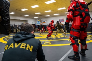 Two people in protective gear practice self-defense techniques on a wrestling mat while dozens of other people in similar, casual and military attire watch from around the room. There is a man crouched in the foreground wearing a hoodie that reads, "Raven."