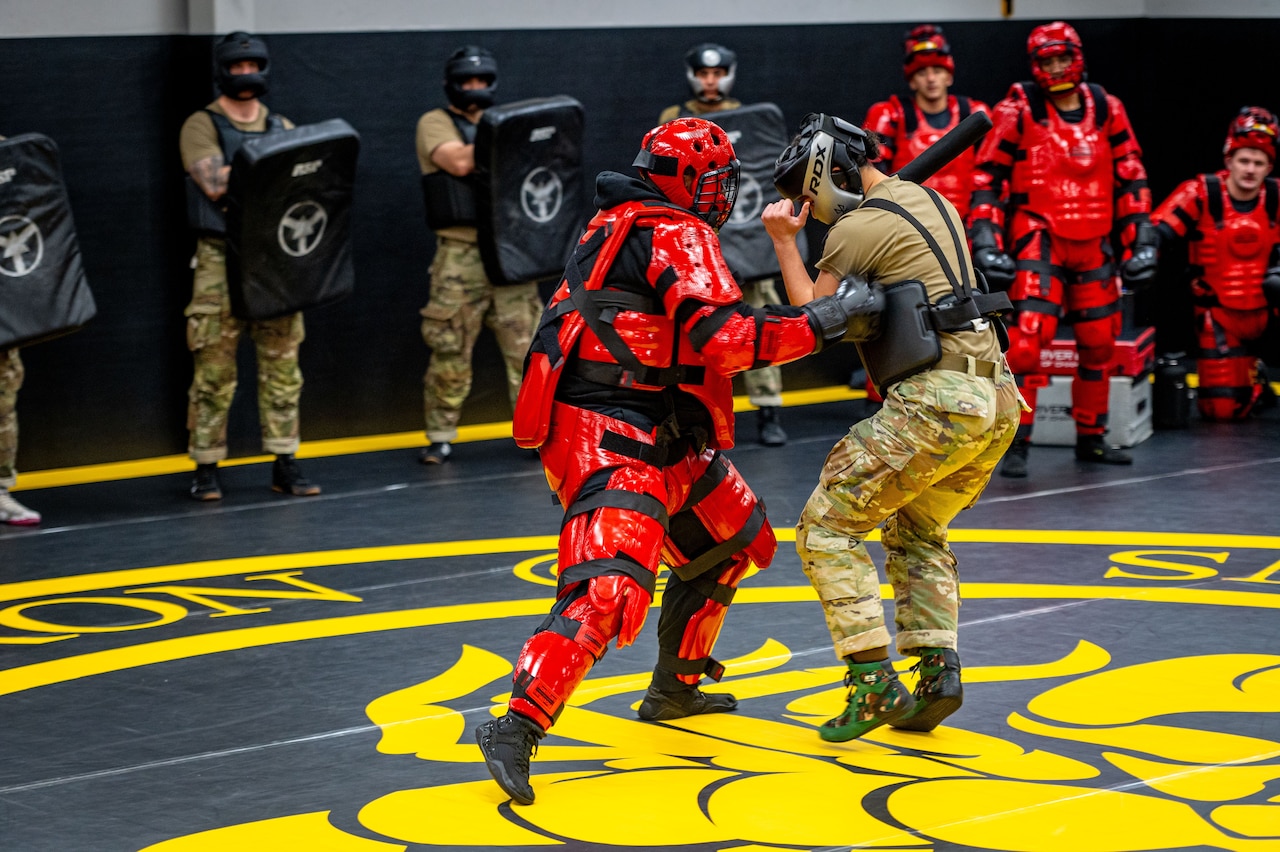 Two people in protective gear practice self-defense techniques on a wrestling mat while seven other people in similar, casual and military attire watch from around the room.