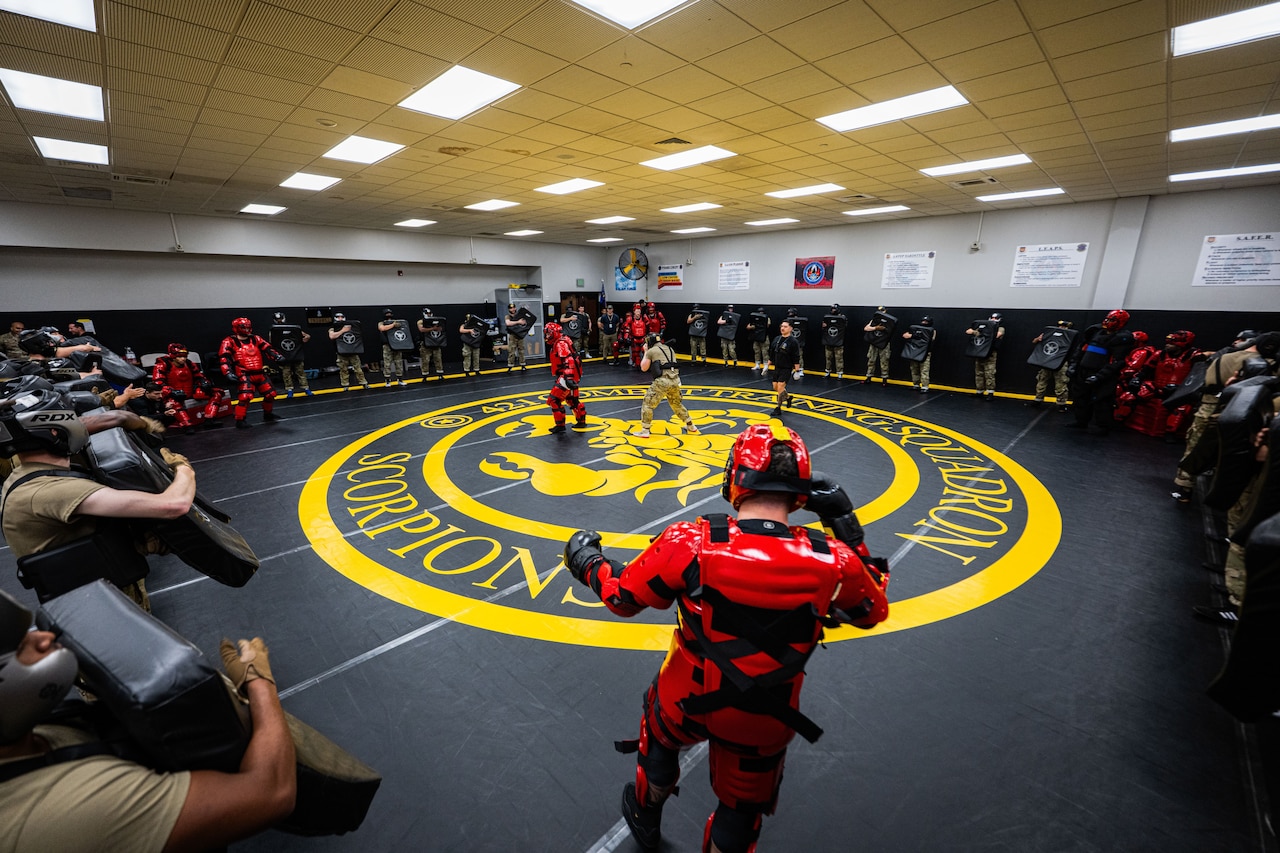 Two people in protective gear practice self-defense techniques on a wrestling mat while dozens of other people in similar, casual and military attire watch from around the room.