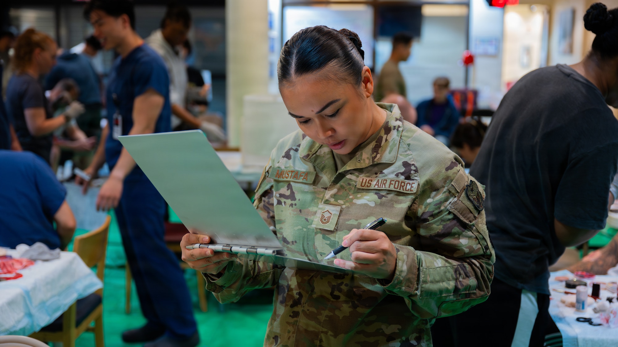 A woman in an OCP camouflage uniform looks down at a clipboard while holding a pen in her hand. The background shows people applying moulage makeup to volunteers.