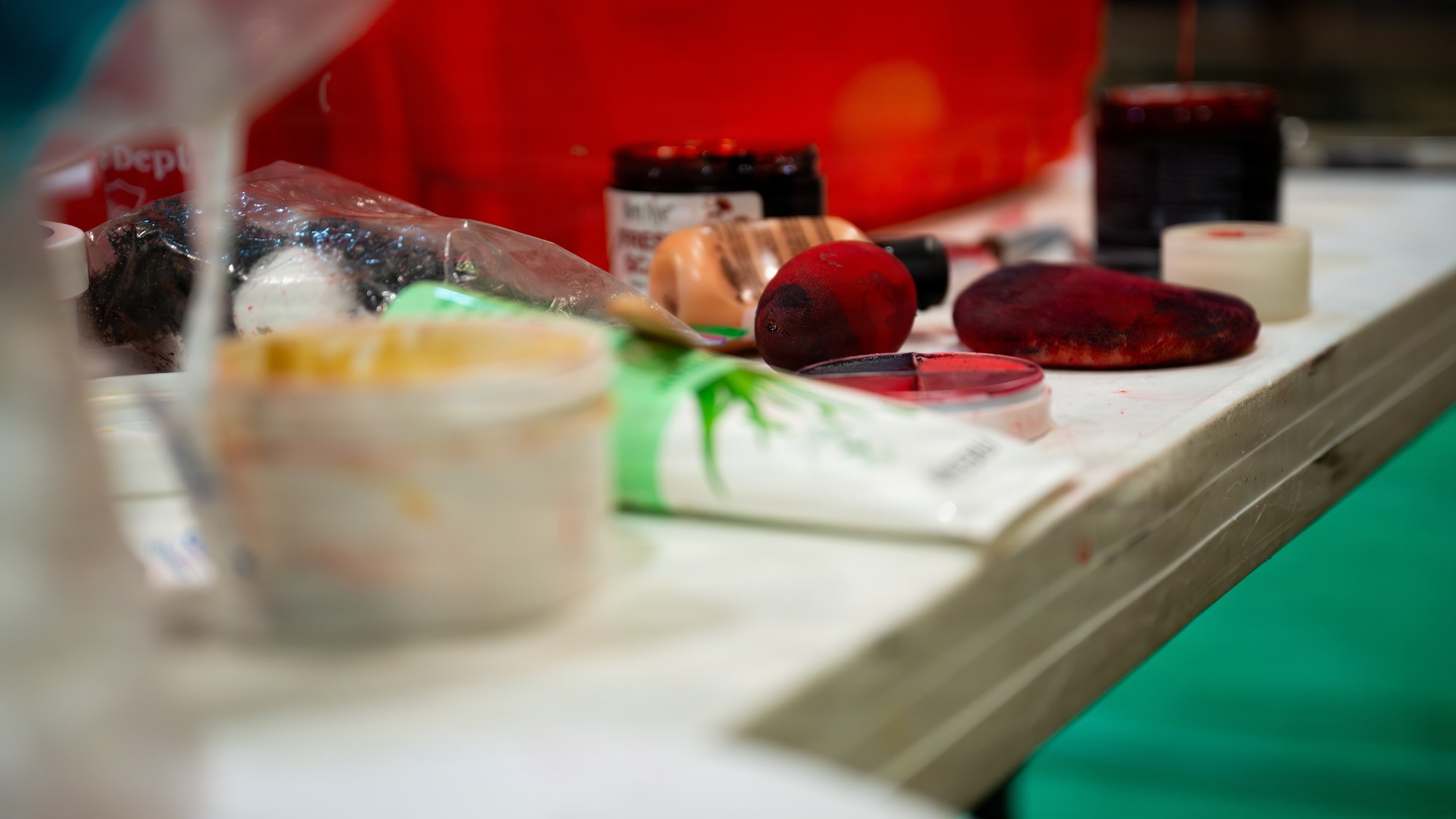 Moulage makeup is spread out on a white table. A red makeup sponge is the focal point and it sits among cream bruise makeup, fake blood, and aloe vera gel