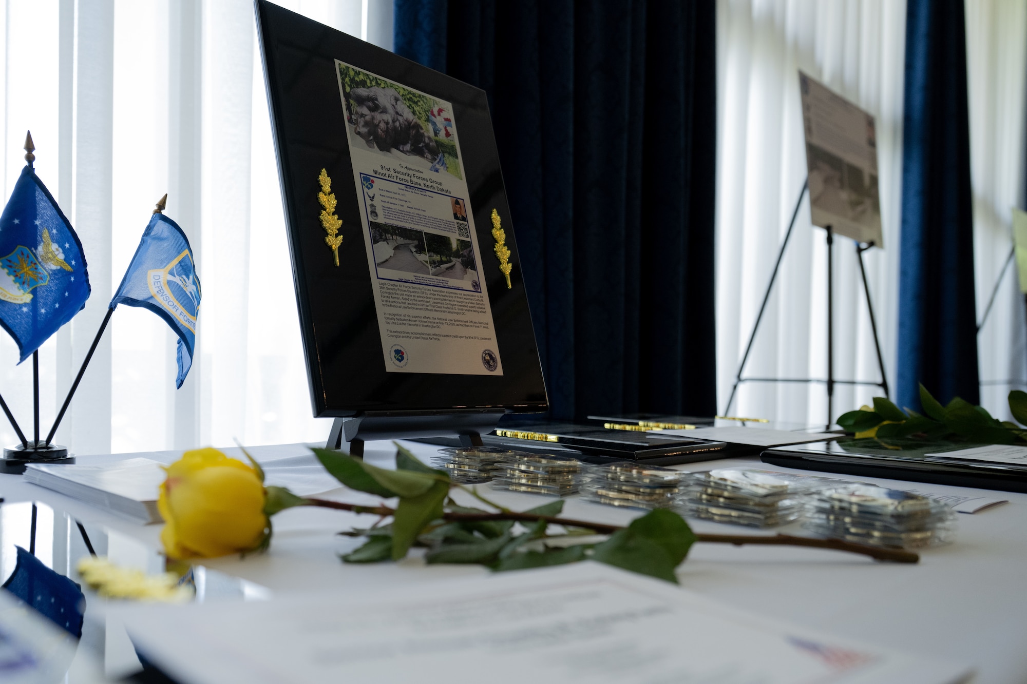 A large placard depicting an Airman's story sits on a white table cloth next to a yellow rose, small Air Force flags, and challenge coins.