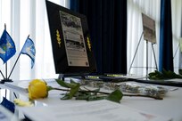 A large placard depicting an Airman's story sits on a white table cloth next to a yellow rose, small Air Force flags, and challenge coins.