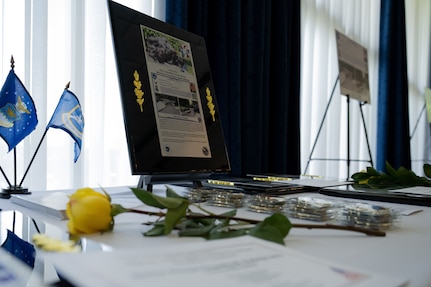 A large placard depicting an Airman's story sits on a white table cloth next to a yellow rose, small Air Force flags, and challenge coins.
