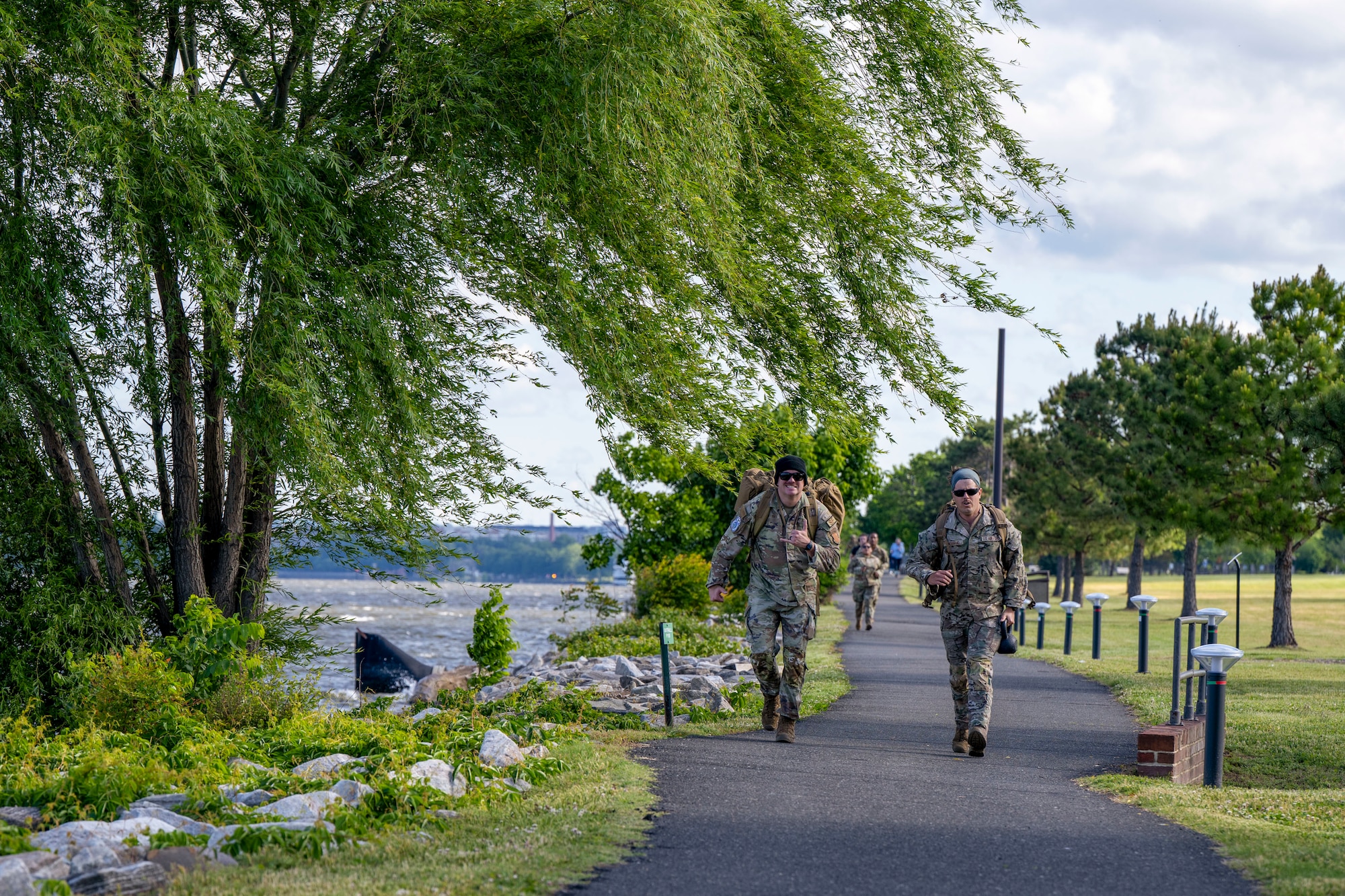 Two Airmen approach marching on a paved path next to the river.