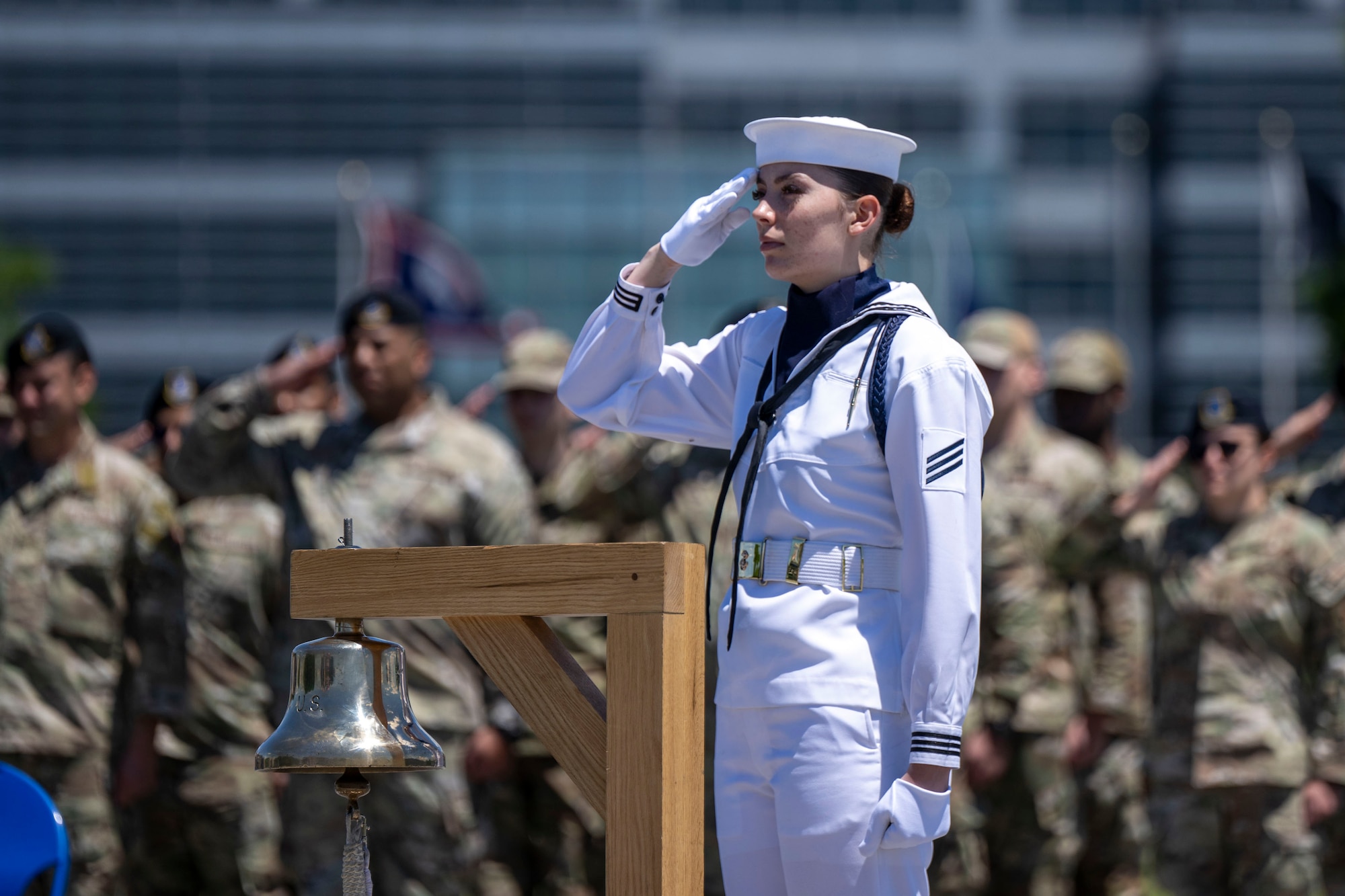 A sailor in dress whites salutes while standing next to a bell.