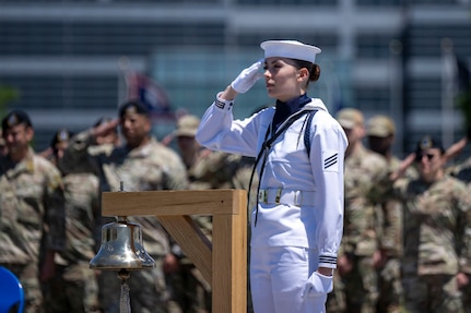 A sailor in dress whites salutes while standing next to a bell.